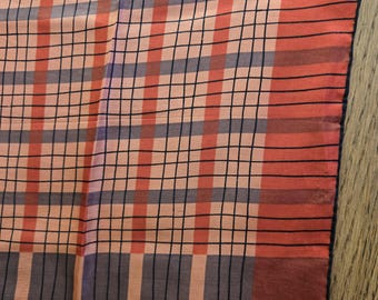 Puede incluir: Primer plano de una bufanda estampada. La bufanda presenta un diseño de cuadrícula con rayas alternas en melocotón, gris y rojo. Finas líneas negras crean el patrón de la cuadrícula. La bufanda es de tejido ligero.