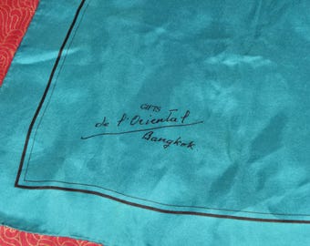 Puede incluir: Pañuelo de seda turquesa con borde negro y el texto "GIFTS de l'Oriental Bangkok" en escritura negra. El pañuelo tiene un brillo sutil y una textura suave. El fondo es una tela roja estampada.