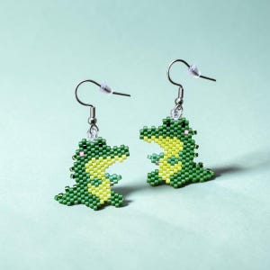 May include: A pair of dangle earrings featuring small crocodile charms crafted from tiny green and yellow beads. Each crocodile has a light green belly, dark green back, and a small pink dot on the cheek. The charms hang from silver-toned metal hooks.