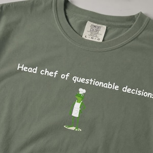 Op de afbeelding: Een saliegroen t-shirt met de witte tekst Head chef of questionable decisions en een afbeelding van een groene kikker met een witte koksmuts en schort. Het t-shirt heeft een Comfort Colors merklabel in de nek met de maat L.