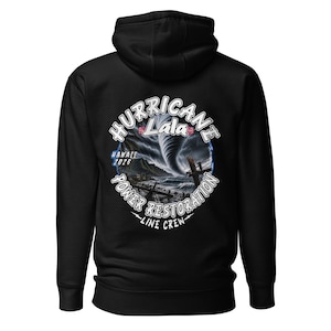 Può includere: Una felpa con cappuccio nera con una grafica sul retro. Il design mostra un tornado sopra un paesaggio costiero con il testo "HURRICANE LALA", "HAWAII 2026", "POWER RESTORATION" e "LINE CREW" stampato in bianco con accenti floreali.