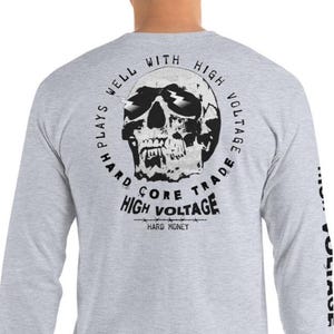 Könnte beinhalten: Hellgraues Langarmshirt mit einem Totenkopf-Motiv auf dem Rücken. Der Totenkopf hat Blitze und ist von dem Text "HARD PLAYS WELL WITH HIGH VOLTAGE CORE TRADE HIGH VOLTAGE HARD MONEY" umgeben. Der Ärmel trägt den Schriftzug "HIGH VOLTAGE".
