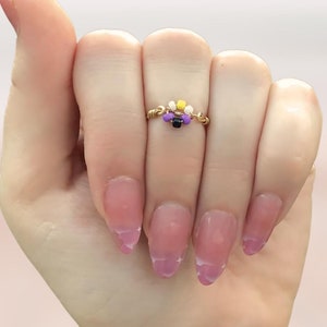 May include: A gold ring with a small beaded design in yellow, white, purple, and black. The ring is worn on the middle finger of a hand with light pink nail polish.
