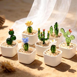 May include: A collection of small, glass cactus sculptures in white square pots. The cacti have various shapes and colors, including green, yellow, and blue, with small, colorful flower accents. The pots are filled with sand-like material.