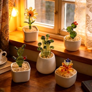 May include: A selection of glass sculptures of succulents and flowers in white ceramic pots. The sculptures include a sunflower, cacti, and other abstract floral designs. The glass is transparent with yellow, green, and red accents. The pots are on a wooden windowsill.