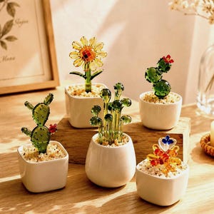 May include: A collection of five miniature glass plants in small white ceramic pots. The set includes a yellow sunflower, a green prickly pear cactus, a tall green succulent, a round green cactus with red flowers, and a multi-colored glass flower.