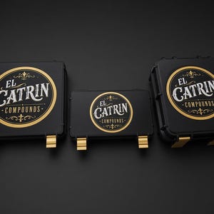 May include: Three black rectangular storage cases with gold-coloured latches. Each case features a circular logo on the lid with the text EL CATRIN COMPOUNDS in white and gold lettering, surrounded by decorative flourishes and a gold border.