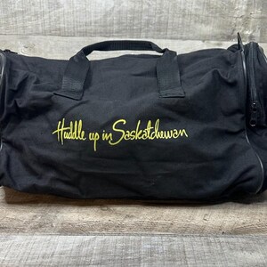 May include: A black duffel bag featuring two top handles and a side zipper compartment. The front is embroidered with the yellow text Huddle up in Saskatchewan. A partial colorful emblem is visible on the left side of the bag.