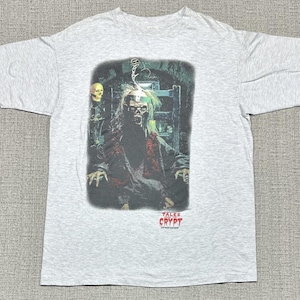 May include: A light gray short-sleeve t-shirt featuring a graphic print of the Crypt Keeper from Tales from the Crypt. The character is depicted in a dark, dungeon-like setting with a skull nearby. The text Tales from the Crypt is printed in red.
