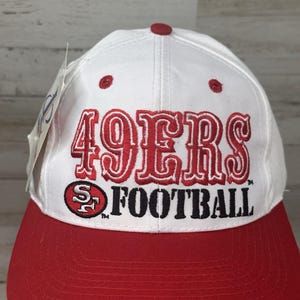 May include: A white and red baseball cap with the text "49ERS FOOTBALL" in red and black. The cap has a red brim and a red button on top. The logo of the San Francisco 49ers is also visible.