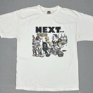 May include: White t-shirt with a cartoon graphic of a football player, an eagle, and other figures. The text "NEXT..." is printed above the graphic.