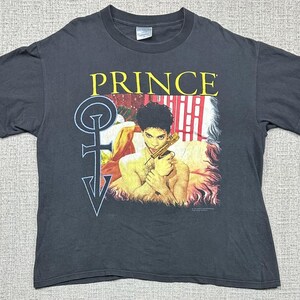 May include: A charcoal gray short-sleeve t-shirt featuring a graphic print of the musician Prince holding a gold-colored object. The text "PRINCE" is printed in yellow above the image, with a large light blue symbol to the left of the figure.