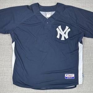 May include: Navy blue baseball jersey with white accents and the New York Yankees logo on the chest. The jersey has a button-down front and short sleeves. A Majestic Authentic Collection label is visible.