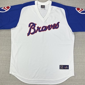 May include: White baseball jersey with blue sleeves and red accents. The word "Braves" is written across the chest in red script with a blue outline. The sleeves have a blue and white design. A small patch is visible near the bottom.