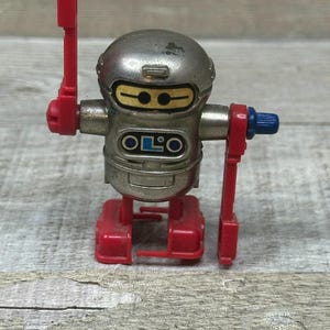 May include: A vintage toy robot with a silver head and body, red feet and arms, and a blue knob. The robot has a rectangular body with the word "OLO" printed on it. The robot's arms are in a raised position, and it has yellow eyes.