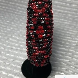 May include: A cylindrical beaded object, primarily red and black, is displayed on a black stand. The beads are tightly arranged, creating a textured surface. The design features a central pattern of red beads.