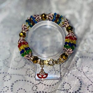 May include: A colorful beaded bracelet with gold accents and a red mask charm. The bracelet features a variety of beads in red, yellow, green, blue, and gold. The charm is a small, red mask with gold detailing, hanging from a gold clasp.