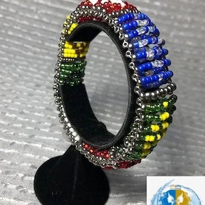 May include: A beaded bracelet with a colorful design. The bracelet features red, white, blue, green, and yellow beads, with silver-colored beads outlining the edges. The bracelet is displayed on a black stand.