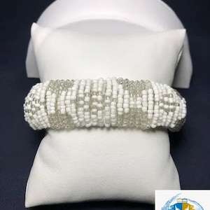 May include: A white beaded bracelet with clear and white beads. The bracelet is displayed on a white pillow and a white curved stand, set against a dark blue background. The bracelet appears to be approximately 1 inch wide.