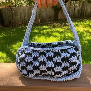 Może przedstawiać: Ręcznie robiona szydełkowa torebka w pepitkę w kolorze czarnym i jasnoniebieskim. Torba ma zaokrąglony kształt z jasnoniebieskim uchwytem. Torba leży na drewnianej powierzchni.