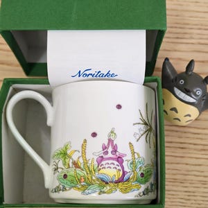 Könnte beinhalten: Weiße Keramiktasse mit einer skurrilen Illustration eines lila-weißen Wesens, umgeben von Pflanzen. Die Tasse befindet sich in einer grünen Schachtel mit dem Markennamen "Noritake". Eine kleine Figur des Wesens befindet sich neben der Tasse.