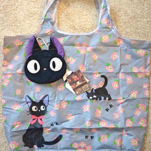 May include: A light blue tote bag with a floral pattern and illustrations of a black cat with a pink bow. Includes a black cat face keychain with purple ears. The bag has a white trim.
