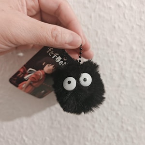 Könnte beinhalten: Ein schwarzer, flauschiger Soot Sprite-Schlüsselanhänger mit großen weißen Augen. Der Schlüsselanhänger ist an einem kleinen Anhänger mit japanischem Text und einer Illustration einer Figur aus dem Film Spirited Away befestigt.
