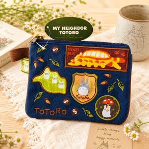 Könnte beinhalten: Eine blaue Canvas-Tasche mit aufgestickten Aufnähern von Charakteren und Szenen aus "Mein Nachbar Totoro". Die Tasche hat einen Reißverschluss, einen braunen Griff und die Wörter "TOTORO" und "NEKO BUS" sind sichtbar.