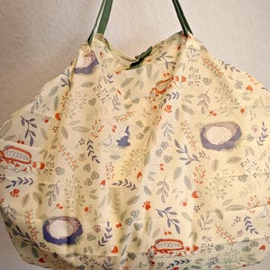 Könnte beinhalten: Eine cremefarbene Tragetasche mit einem skurrilen Waldmuster mit Nestern, Waldtieren und Blättern in Blau-, Orange- und Grüntönen. Die Tasche hat grüne Träger.