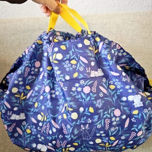Könnte beinhalten: Ein marineblauer Kordelzugbeutel mit einem floralen Muster aus weißen Katzen, rosa und gelben Blumen und grünen Blättern. Die Tasche hat leuchtend gelbe Griffe. Die Tasche wird von einer Hand gehalten.