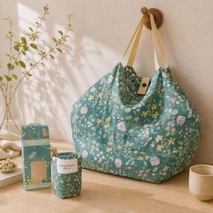 Könnte beinhalten: Eine türkisfarbene Einkaufstasche mit floralem Muster, die an einem Holzhaken hängt. Die Tasche hat gelbe Träger und einen Knopfverschluss. Eine passende aufgerollte Tasche und eine Schachtel daneben, beide mit dem Text "Shupatto" und "Shopping Bag".