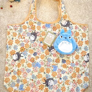 Op de afbeelding: Een totebag met bloemenpatroon met een lichtbeige achtergrond, oranje, roze en blauwe bloemen en stripfiguren. Een blauw pluche karakter en een klein label staan op de tas. De tas heeft een bruine rand en handvatten.