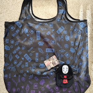 Könnte beinhalten: Schwarze Tragetasche mit einem sich wiederholenden Muster aus blauen und lila japanischen Illustrationen. Eine kleine schwarze Plüschtasche mit einem weißgesichtigen Charakter und einem roten Akzent hängt an der Tragetasche. Ein kleines Etikett mit Zeichentrickfiguren ist ebenfalls angebracht.
