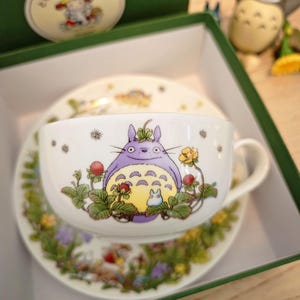 Könnte beinhalten: Weißes Keramik-Teeservice mit lila Totoro-Motiv und floralen Details. Das Set wird in einer grünen Box mit passendem Deckel und einer Totoro-Figur präsentiert. Dekorationen auf Tasse und Untertasse.
