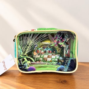 Könnte beinhalten: Eine rechteckige grüne Tasche mit einer transparenten Vorderseite, die eine detaillierte Miniatur-Schlafzimmerszene zeigt. Das Innere umfasst ein Bett mit einer grün-weiß karierten Decke, ein Bücherregal und dekorative Elemente.
