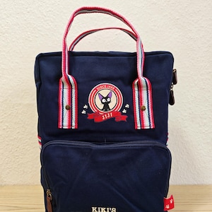 Könnte beinhalten: Marineblauer Rucksack mit einer Vordertasche und gestreiften Griffen. Der Rucksack hat einen gestickten Aufnäher mit einer schwarzen Katze und dem Text "JIJI". Der Text "KIKI'S DELIVERY SERVICE" ist auf der Vordertasche aufgedruckt.