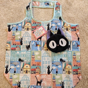 Könnte beinhalten: Eine wiederverwendbare Einkaufstasche mit einem farbenfrohen Stadtlandschaftsmuster mit Illustrationen von schwarzen Katzen. Die Tasche hat einen passenden schwarzen Katzenförmigen Beutel. Ein kleines Etikett mit Illustrationen ist ebenfalls sichtbar.
