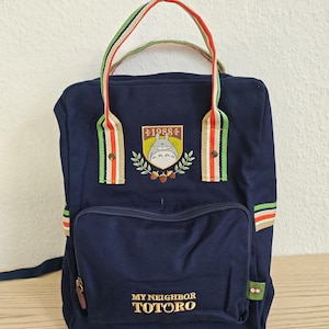 Könnte beinhalten: Marineblauer Rucksack mit dem goldenen Schriftzug "MY NEIGHBOR TOTORO". Er hat eine Vordertasche und einen Aufnäher mit dem Jahr "1988" und einer Totoro-Figur. Die Griffe sind grün, rot und weiß gestreift.