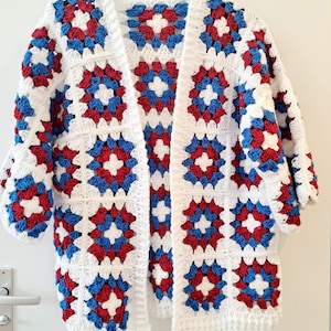 Puede incluir: Un cárdigan de crochet blanco con frente abierto y diseño de patrón cuadrado. Los cuadrados presentan patrones florales rojos y azules. El cárdigan está colgado de una percha negra sobre un fondo blanco.