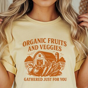 May include: Yellow t-shirt with the text "ORGANIC FRUITS AND VEGGIES" in brown. The graphic includes a barn, fruits, and vegetables. The phrase "GATHERED JUST FOR YOU" is below the graphic. The model is wearing blue denim shorts.