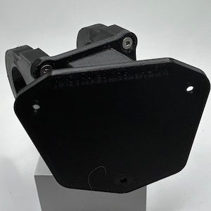 May include: Black plastic 3D printed part with two visible screws. The part has a rounded, curved shape and a flat, rectangular base.