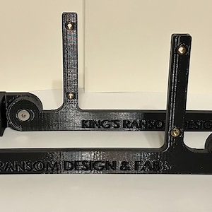May include: Two black 3D printed brackets with the text "KING'S RANSOM DESIGN & FAB" on the bottom. Each bracket has a round, black, adjustable clamp on the side.