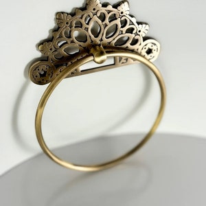 May include: A gold-toned metal towel ring with a decorative, ornate, floral design. The ring is circular and has a delicate, intricate pattern.