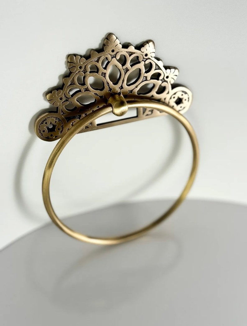 May include: A gold-toned metal towel ring with a decorative, ornate, floral design. The ring is circular and has a delicate, intricate pattern.