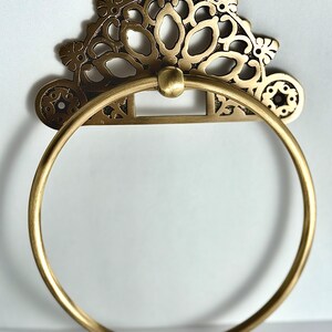 May include: A gold-toned metal towel ring with an ornate, decorative design. The ring is circular and has a decorative, floral-inspired design.