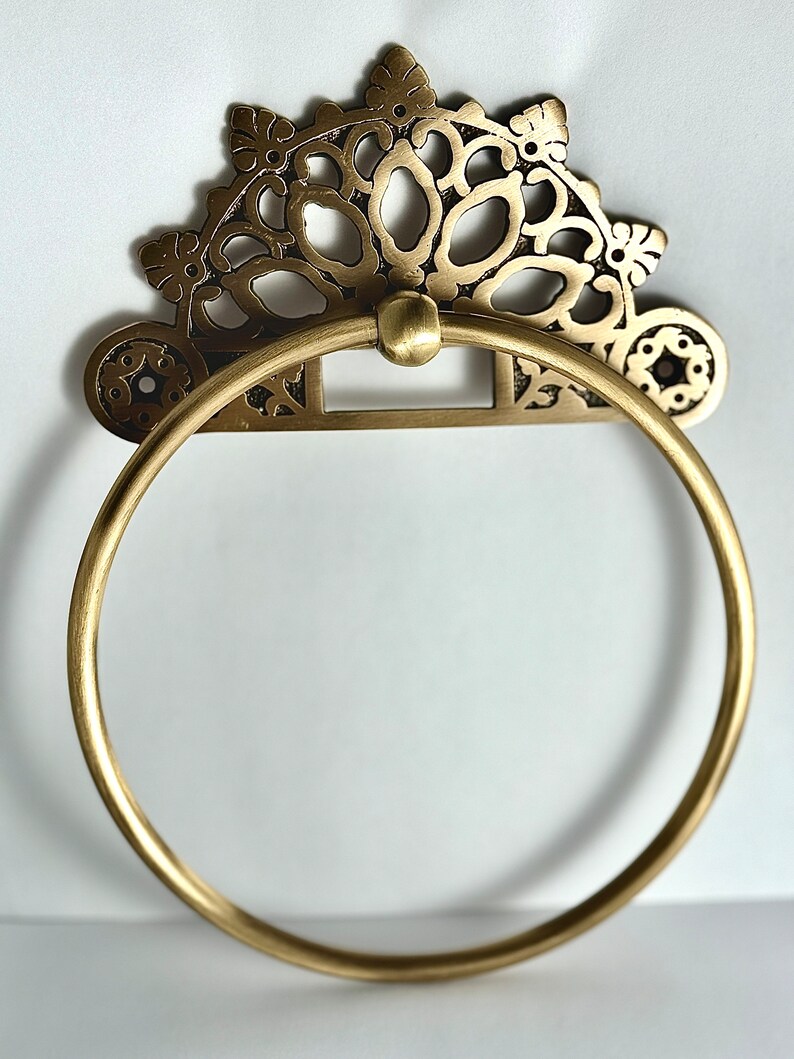 May include: A gold-toned metal towel ring with an ornate, decorative design. The ring is circular and has a decorative, floral-inspired design.