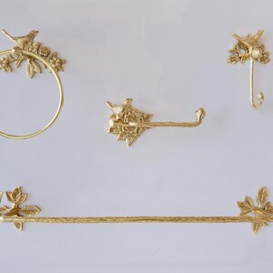 May include: A set of gold-colored bathroom hardware, including a towel ring, toilet paper holder, hook, and towel bar. Each piece features a bird and floral design. The hardware is mounted on a white wall.
