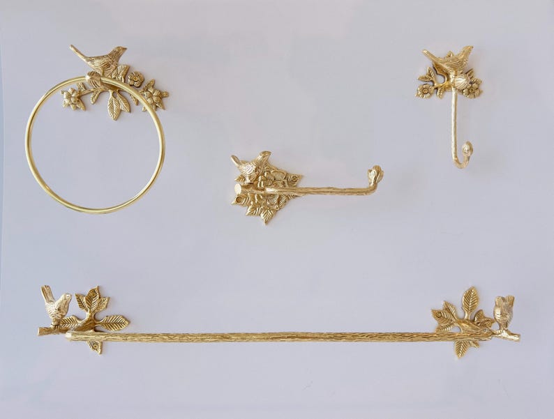 May include: A set of gold-colored bathroom hardware, including a towel ring, toilet paper holder, hook, and towel bar. Each piece features a bird and floral design. The hardware is mounted on a white wall.