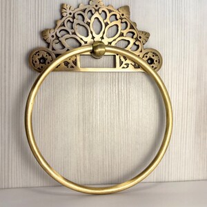 May include: A gold-toned metal towel ring with an ornate, decorative top. The ring is circular and has a smooth, polished finish.