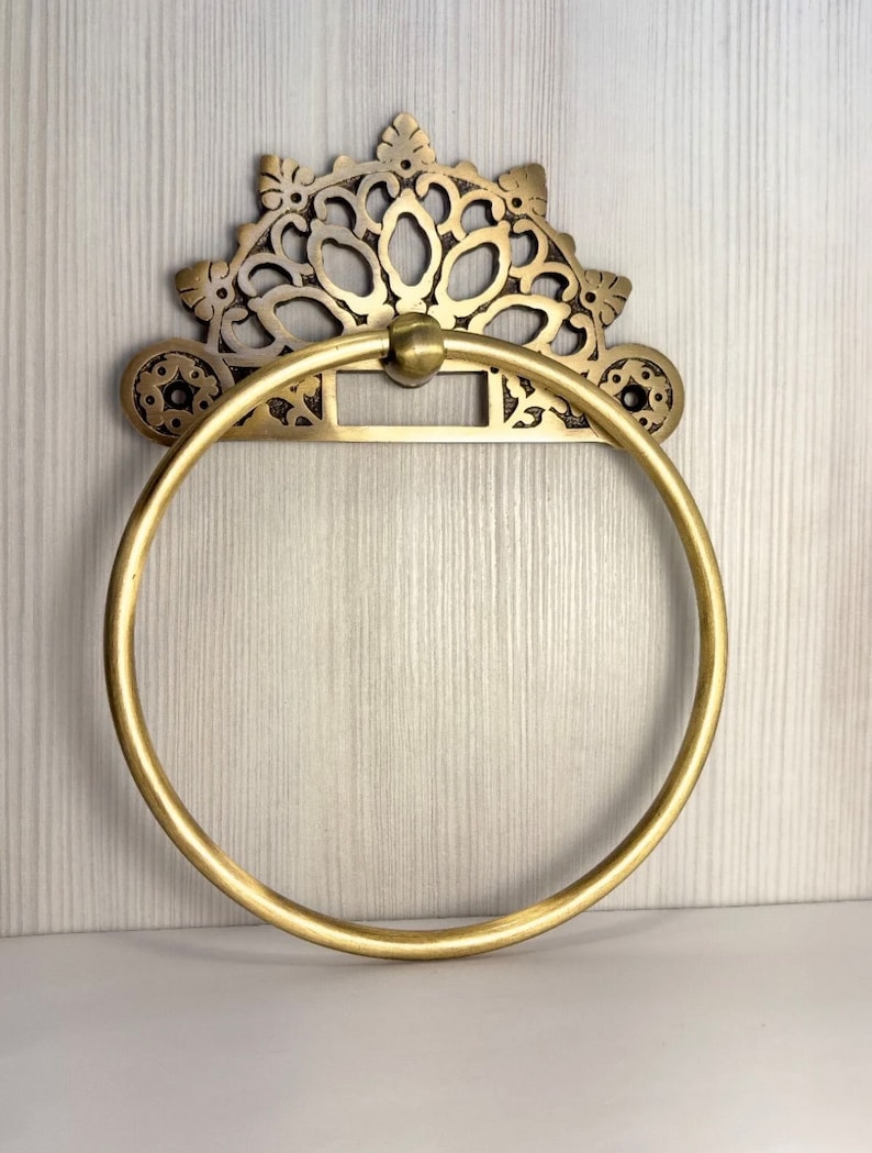 May include: A gold-toned metal towel ring with an ornate, decorative top. The ring is circular and has a smooth, polished finish.